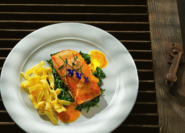 Ein angerichteter Teller mit gebratenem Lachsfilet, grünem Gemüse, gelben Komponenten und Spiezenblumen.