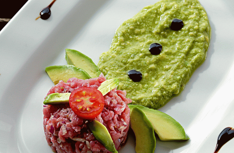 Ein kunstvoll angerichtetes Gericht mit Avocado-Dip, rotem Hackfleisch und Tomaten auf einem weißen Teller.